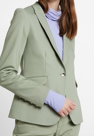 Vrouw draagt een lichtgroene, op maat gemaakte blazer over een lavendelkleurige coltrui, met bijpassende lichtgroene broek, poseert met één hand bij de blazer.