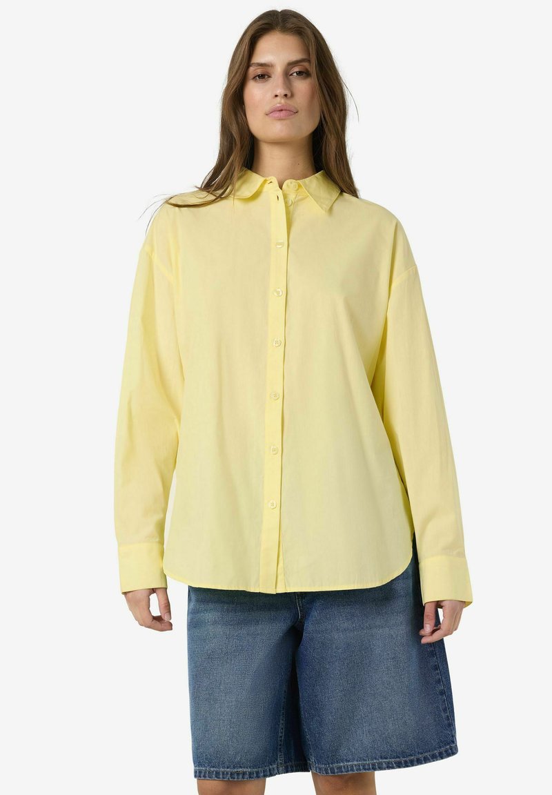 Femeie purtând o cămașă galben deschis, largă, cu nasturi și mâneci lungi, și pantaloni scurți din denim cu spălare medie, stând în picioare pe fundal alb.