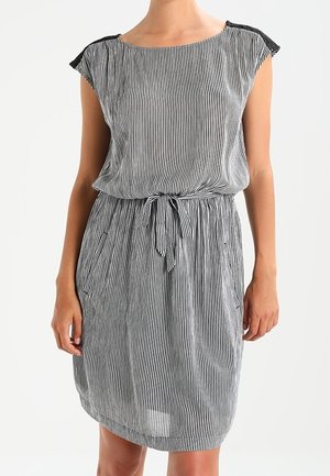 Kvinde iført en knælængde, ærmeløs kjole med sorte og hvide vertikale striber, et bundet bælte i taljen og detalje ved skulderen.