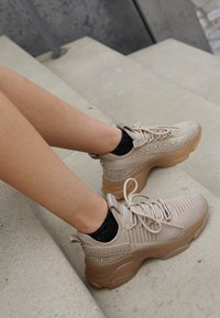Beige knit sneakers with textured pattern, sparkling accents, and a transparent gum rubber sole, paired with black glitter socks.