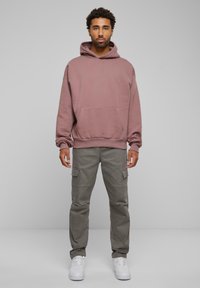 Sudadera de gran tamaño en color rosa polvo con bolsillo frontal, combinada con pantalones cargo grises claros que cuentan con bolsillos laterales, y zapatillas blancas.