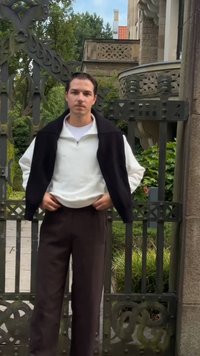 Fekete félzipzáras mellény egy fehér felső felett, sötétbarna nadrággal párosítva. Egy dekoratív fémkapu és zöld növényzet előtt áll.