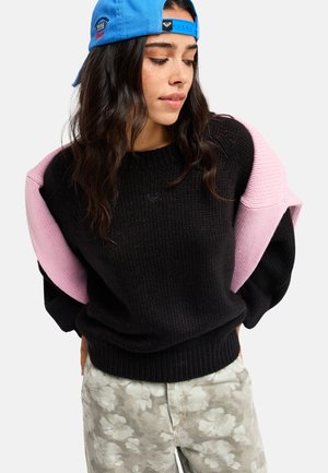 Junge Frau, die eine blaue rückwärts getragene Kappe, einen schwarzen Pullover, einen rosa Pullover über die Schultern gelegt und Hose mit Blumenmuster trägt und nach unten schaut.