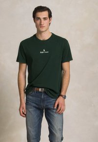 Mlad moški stoji oblečen v temno zeleno majico Ralph Lauren, modre kavbojke, rjav pas in ročno uro pred nevtralno ozadje.