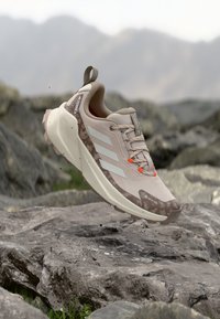 Chaussure de randonnée beige avec une tige texturée, des bandes blanches et des accents orange. Dispose d'une semelle amortie et d'une semelle extérieure robuste pour une meilleure traction.