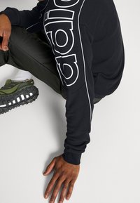 Sudadera negra con detalle en logo blanco, combinada con pantalones verde oliva y zapatillas negras resistentes con acentos blancos. Textura de tela suave.