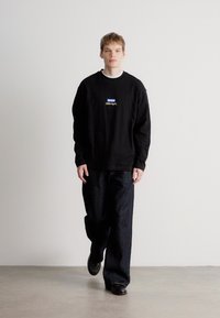 Μαύρη φούτερ μπλούζα με μακριά μανίκια και λογότυπο στο στήθος, σε συνδυασμό με σκούρες μπλε φαρδιές παντελόνες και μαύρα παπούτσια σε ουδέτερο φόντο.