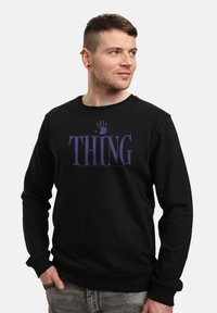 Uomo con capelli corti che indossa una felpa nera con il testo viola "THING" e un design di impronta di mano, che guarda di lato, con una mano in tasca.