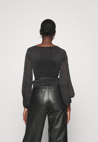 Svart långärmad topp med figurnära passform och rynkdetaljer vid midjan, tillverkad av glittrande tyg. Kombinerad med högmidjade läderbyxor.