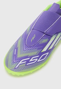Chaussure de football violette avec un design épuré, une surface texturée agrémentée d'accents verts et une sangle Velcro. Branding notable F50 et Sprintframe.