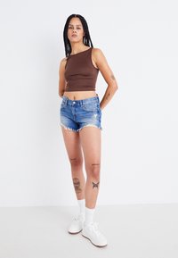 Barna ujjú crop top, magas derekú, koptatott kék farmer sort, rojtos szegéllyel, fehér sportcipő és fehér bokazokni, lábon látható tetoválások.
