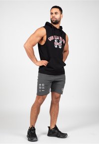 Zwarte mouwloze hoodie met witte en rode graphics, gecombineerd met grijze shorts met verschillende bedrukte logo's. Zwarte sportschoenen.