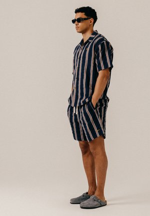 Homme portant des lunettes de soleil noires, une chemise à manches courtes rayée bleu marine et beige et un short assorti, debout les mains dans les poches et chaussures grises à enfiler.