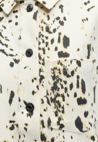 Κοντινή λήψη από κρεμ ύφασμα με μαύρα και ανοιχτό καφέ ακανόνιστα σημεία, με μαύρα κουμπιά κράτησης και τσέπη στο στήθος.