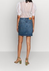 Žena v průhledné bílé blůze s balónovými rukávy, modré džínsové minisukni a olivově zelených sandálech na nízkém podpatku, otočená zády.