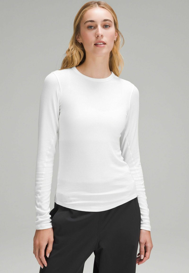 Maglietta bianca a maniche lunghe realizzata in materiale liscio e leggero, con scollatura a girocollo e design aderente, abbinata a pantaloni neri.