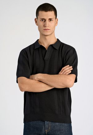 Black knitted polo shirt with short sleeves, featuring vertical ribbed texture and a three-button placket, paired with dark denim jeans.