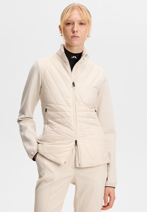 Kvinde med blondt hår iført en cremefarvet quiltet jakke over en sort top, hænderne i lommerne, foran en ensfarvet lys grå baggrund.