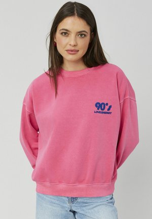 Jeune femme aux longs cheveux foncés portant un sweat-shirt rose avec le texte "90's LOVE ENERGY" et un jean bleu clair, devant un fond uni.