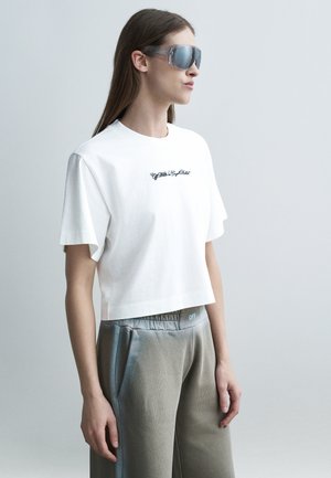 Γυναίκα με πολύ μεγάλα γυαλιά ηλίου με καθρέφτη, λευκό κοντό T-shirt με μικρό μαύρο κείμενο και άνετο γκρι παντελόνι με ελαστική μέση.