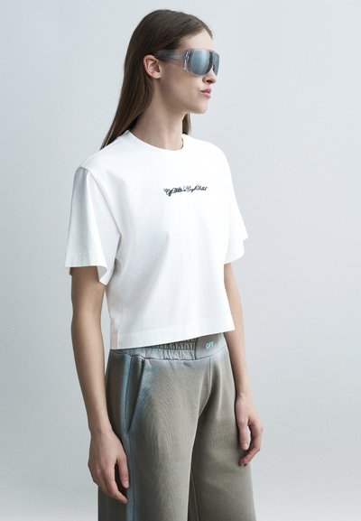 Frau trägt übergroße verspiegelte Sonnenbrille, weißes bauchfreies T-Shirt mit kleinem schwarzem Text und lockere graue Hosen mit elastischem Bund.