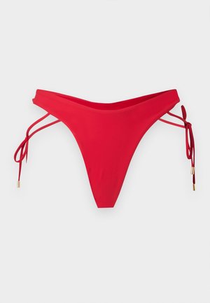 Γυναίκα όρθια ξυπόλητη, φοράει καφέ μπικίνι με μπανέλα και κόκκινo μπικίνι κάτω μέρος με κορδόνια στα πλάγια, σε λευκό φόντο.