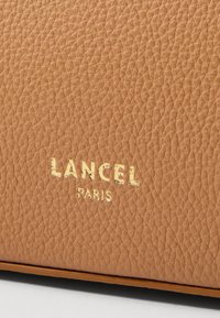 Texture in pelle color cuoio con una finitura a ciottoli, caratterizzata dal logo dorato in rilievo "LANCEL" e "PARIS" vicino al bordo inferiore.