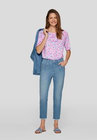 Pink and blue patterned short-sleeve top, light blue high-waisted jeans with embellished pockets, and blue slip-on sandals. Denim jacket draped over shoulder.