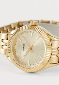 Montre Timex dorée avec un cadran texturé, un cadran lisse, des index horaires et un bracelet en métal. Boîtier rond avec une couronne à 14 heures.