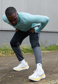 Ténis desportivos em branco com detalhes em laranja-amarelo, apresentando uma parte superior em malha, sola acolchoada e detalhes texturizados. Usados com calças desportivas cinzentas.