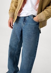 Osoba oblečená v modrých džínách, béžové bundě a bílé košili, s jednou rukou v kapse a druhou povolenou u těla.