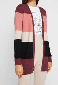 Kardigan med blockfärger: vinröd, rosa, beige och svart. Mjuk textur, öppen framsida, ribbade ärmslut och detalj i nederkant. Visar en avslappnad stil.