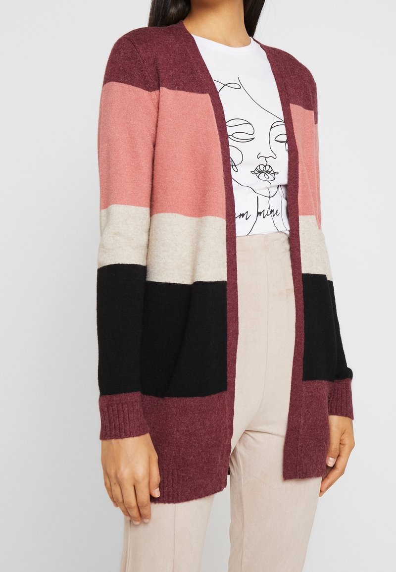 Kardigan med blockfärger: vinröd, rosa, beige och svart. Mjuk textur, öppen framsida, ribbade ärmslut och detalj i nederkant. Visar en avslappnad stil.