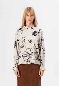 Donna bionda che indossa una blusa lunga beige con stampa floreale nera e un collo arricciato alto, abbinata a una gonna marrone.