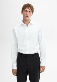 Weiße Bluse mit spitzen Kragen, langen Ärmeln und durchgehender Knopfleiste; getragen mit schwarzen Hosen, schlicht im Design.