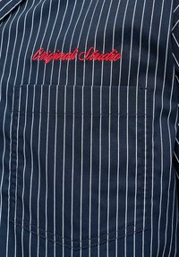 Tissu bleu marine avec fines rayures verticales blanches, une poche poitrine et un texte brodé rouge indiquant "Original Studio."