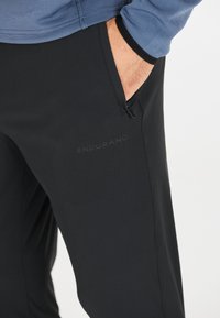 Zwarte sportbroek met een gladde textuur, voorzien van een ritszak en het woord "ENDURANCE" subtiel op de zijkant gebosseleerd.