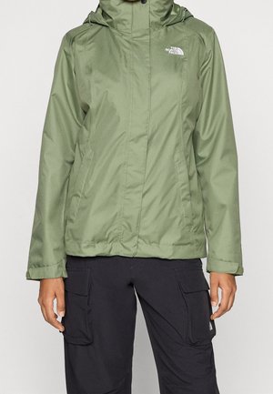 Osoba ubrana w oliwkową kurtkę North Face z kapturem i czarne spodnie cargo, stojąca na tle jednolitego jasnego tła.