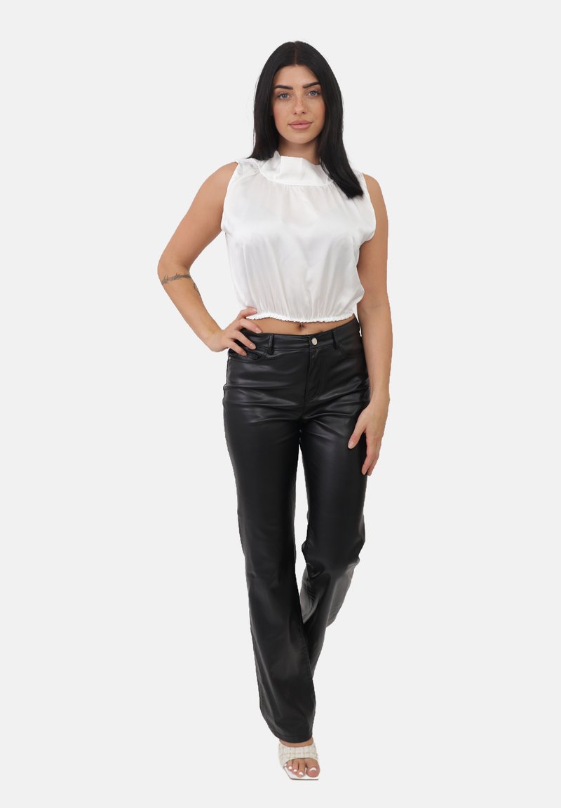 Donna in piedi con la mano sui fianchi, che indossa un top corto senza maniche in satin bianco, pantaloni in pelle nera e sandali con tacco bianchi.