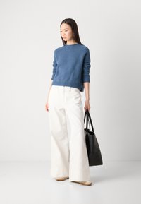 Blauwe wollen trui met een ronde hals, gecombineerd met witte wijde broekspijpen. Het model houdt een zwarte tote bag vast, wat het contrast en de minimalistische uitstraling benadrukt.