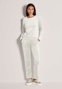 Frau steht mit den Händen in den Taschen, trägt einen hellgrauen gemusterten Pullover, passende Hosen und Sneakers mit Schlangenledermuster auf Plateausohle.