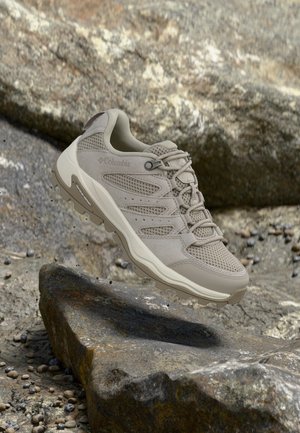 Licht beige Columbia wandelschoen met mesh panelen en robuuste zool zweeft boven rotsachtig terrein.