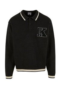 Černé pletené polo s bílým proužkovaným límcem, na prsou má dekorativní nášivku "K" se hvězdami. Hranaté manžety a lem.