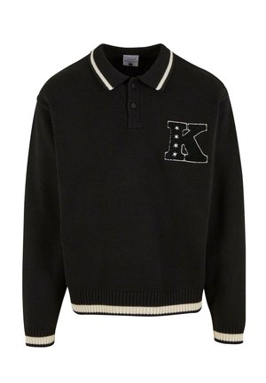Melnā adītā polo krekls ar baltu svītru apkaklē, ar dekoratīvu "K" uzplecu, kas rotāts ar zvaigznēm krūtīs. Ribota aproce un apakša.