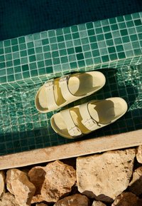 Yellow sandals with double straps placed on the edge of a green-tiled pool, sunlight reflecting on water, and rocks below the pool edge.