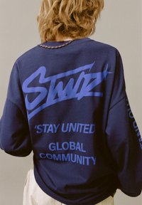 Hanorac bleumarin cu croială oversize, având text grafic albastru: „STAY UNITED” și „GLOBAL COMMUNITY” pe spate și pe mânecă. Textură moale.