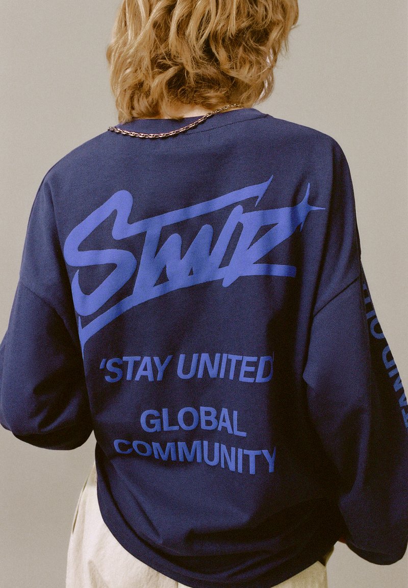 Hanorac bleumarin cu croială oversize, având text grafic albastru: „STAY UNITED” și „GLOBAL COMMUNITY” pe spate și pe mânecă. Textură moale.