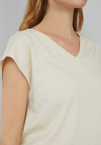 Vrouw in een lichtcrème V-hals shirt met korte mouwen, waarbij nek, schouder en bovenarm zichtbaar zijn tegen een effen grijze achtergrond.