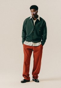 Mann iført runde briller, mørk grønn knappet cardigan over stripete skjorte, røde bukser og svarte loafers, som står med hendene i lommene.