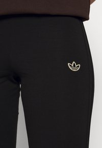Musta legginsit, joissa on pehmeä ja joustava kangas. Ominaisuuksiin kuuluu pieni, brodeerattu kultainen Adidas-logo vyötärön lähellä.